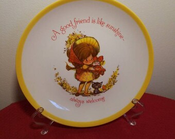 Puede incluir: Un plato decorativo con borde amarillo y centro blanco. El plato presenta una ilustración de dibujos animados de una niña sosteniendo una muñeca y un gato negro. El texto "A good friend is like sunshine - always welcome" está escrito en el plato.