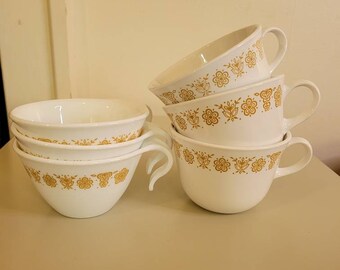 May include: A stack of white ceramic soup bowls and mugs with a gold floral pattern. The mugs have large handles and are stacked on top of each other. The bowls are stacked on the left side of the image.