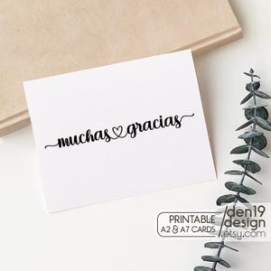 Op de afbeelding: Een witte kaart met de woorden "muchas gracias" in elegant zwart schrift, met een hartsymbool tussen de woorden. De kaart ligt op een neutrale achtergrond met een takje groen.