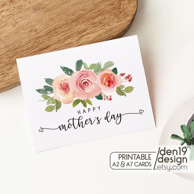 May include: A white Mother's Day card with a watercolor floral design. The card features pink and peach roses with green leaves. The words "Happy Mother's Day" are written in cursive. The card is on a wooden surface.