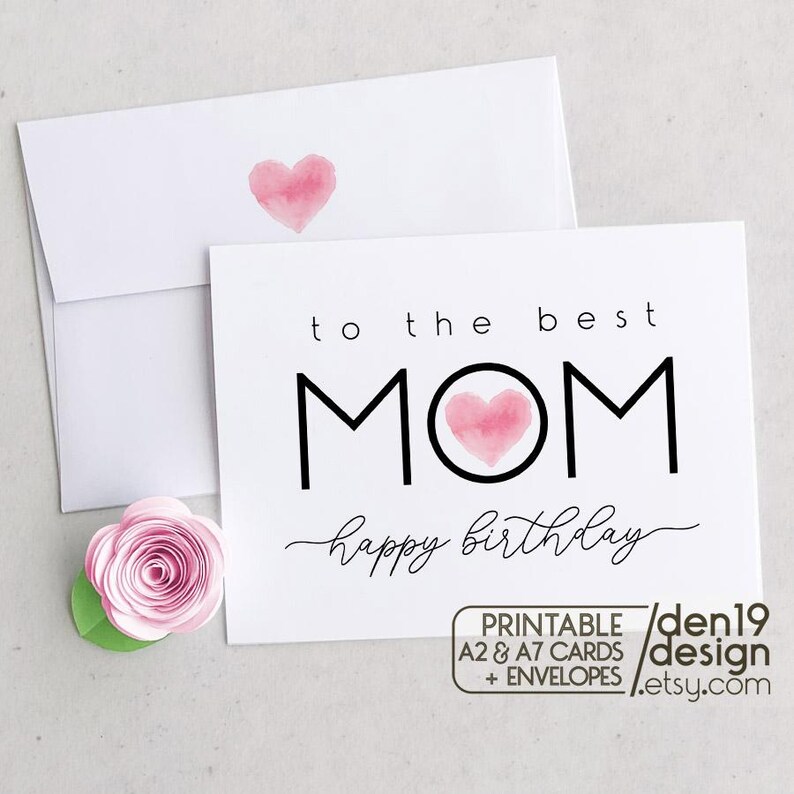 May include: A white greeting card and envelope set with a watercolor heart design. The card reads "to the best MOM happy birthday" in black script and block letters. A pink paper flower is next to the card.