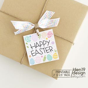 Puede incluir: Una etiqueta de regalo cuadrada de Pascua con las palabras "Happy Easter" e ilustraciones de huevos coloridos. La etiqueta está atada a un paquete marrón envuelto con un lazo de lunares. La etiqueta mide 5.08cm & 6.35cm y es un diseño imprimible.