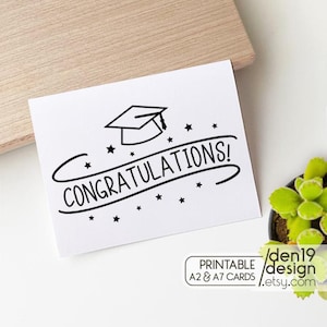 May include: A white graduation card with the word "CONGRATULATIONS!" in a handwritten font, along with a graduation cap and stars. The card is on a wooden surface, with a small plant in the background.