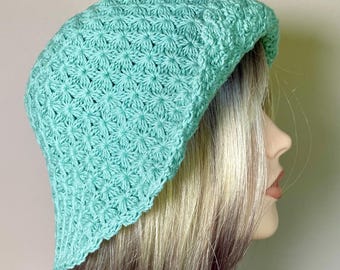 May include: A light mint green crochet bucket hat featuring a repeating star-like stitch pattern throughout the crown and a slightly flared brim. The hat is constructed with a textured yarn finish and maintains a consistent color tone.