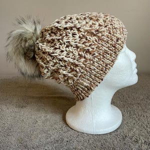 May include: A hand-knitted beanie hat in shades of cream, brown, and tan, topped with a large, fluffy faux fur pom-pom. The hat is displayed on a white mannequin head, showcasing its textured knit design. A cozy winter accessory.