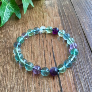 May include: A green, purple, and blue fluorite gemstone bracelet with round beads. The bracelet is on a wooden surface.