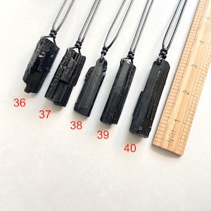 Peut inclure: Cinq pendentifs en tourmaline noire sur cordons noirs, &eacute;tiquet&eacute;s de 36 &agrave; 40, dispos&eacute;s &agrave; c&ocirc;t&eacute; d'une r&egrave;gle en bois gradu&eacute;e en pouces. Chaque pierre brute et verticale pr&eacute;sente une forme allong&eacute;e, textur&eacute;e et unique.