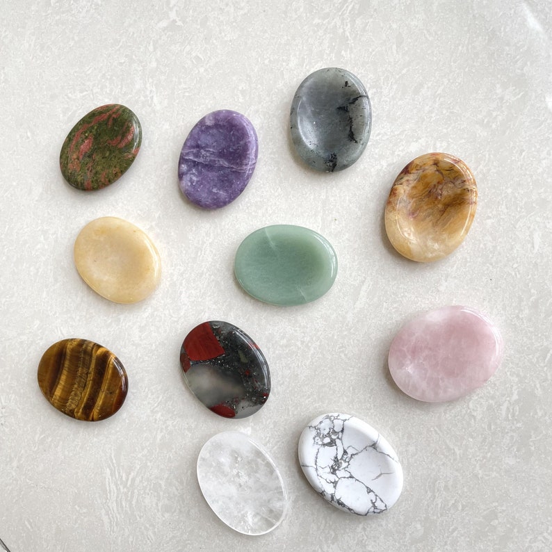 K&ouml;nnte beinhalten: Sortiment von 10 glatten, polierten ovalen Steinen in verschiedenen Farben, darunter Gr&uuml;n, Lila, Rosa, Wei&szlig;, Braun und Schwarz. Die Steine sind in einem Kreis auf einer wei&szlig;en Oberfl&auml;che angeordnet.