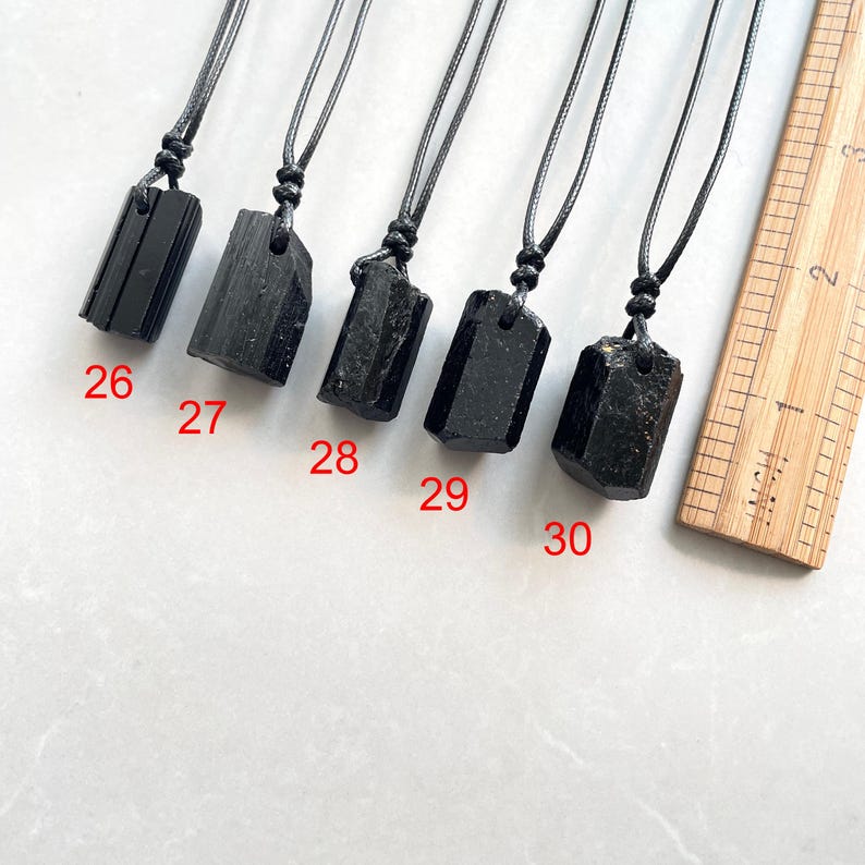 Peut inclure: Cinq pendentifs en cristal brut noir sur des colliers en cordon noir, &eacute;tiquet&eacute;s de 26 &agrave; 30, dispos&eacute;s &agrave; c&ocirc;t&eacute; d'une r&egrave;gle en bois affichant des mesures en centim&egrave;tres. Chaque pendentif pr&eacute;sente une forme de pierre brute unique.