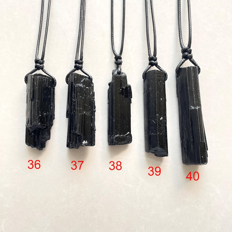 Peut inclure: Cinq pendentifs en tourmaline noire suspendus &agrave; des colliers en cordon noir. Chaque pierre brute et verticale pr&eacute;sente une forme et une texture uniques. Les pendentifs sont marqu&eacute;s des chiffres rouges 36, 37, 38, 39 et 40, identifiant chaque pi&egrave;ce.