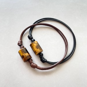 Könnte beinhalten: Zwei Armbänder mit braunen und schwarzen Schnüren und Tigeraugenperlen.