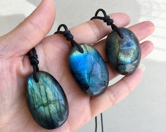 May include: Three oval-shaped labradorite pendants suspended on black cord necklaces. Each polished gemstone displays iridescent flashes of blue, green, and gold light against a dark, semi-translucent base with visible natural internal fractures.