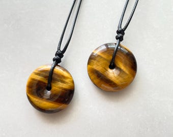 May include: Two tiger's eye stone pendants with black cords. The pendants are circular, with a polished, banded appearance in shades of brown and gold. The cords are knotted at the top.