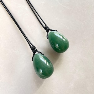 Op de afbeelding: Twee groene jade-eivormige hangers aan zwarte koorden. De hangers zijn glad en gepolijst.