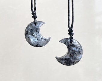 May include: Two crescent moon-shaped pendants. Each pendant is made of a gray and black speckled stone, suspended from a black cord. The pendants are displayed against a neutral background, highlighting their unique shape and texture.
