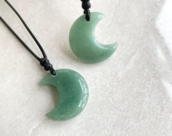 May include: Two green crescent moon pendants on black cords. The pendants are made of a smooth, polished stone. The cords are knotted at the top, suggesting an adjustable length. The background is a light, textured surface.