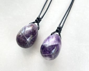 Puede incluir: Dos colgantes de amatista en forma de huevo con cordones negros. Las piedras pulidas muestran una mezcla de tonos morados y blancos. Los cordones están anudados en la parte superior, lo que sugiere que están diseñados para usarse como collares.