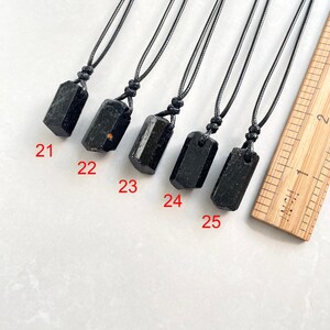 Peut inclure: Cinq pendentifs en cristal brut noir sur des colliers en cordon noir, num&eacute;rot&eacute;s de 21 &agrave; 25, dispos&eacute;s &agrave; c&ocirc;t&eacute; d'une r&egrave;gle en bois affichant des mesures en pouces. Chaque pendentif pr&eacute;sente une pierre sombre, brute et allong&eacute;e.