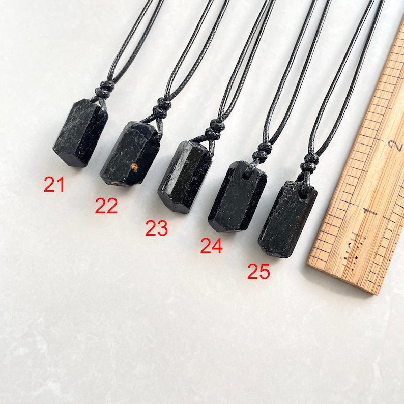 Peut inclure: Cinq pendentifs en cristal brut noir sur des colliers en cordon noir, num&eacute;rot&eacute;s de 21 &agrave; 25, dispos&eacute;s &agrave; c&ocirc;t&eacute; d'une r&egrave;gle en bois affichant des mesures en pouces. Chaque pendentif pr&eacute;sente une pierre sombre, brute et allong&eacute;e.