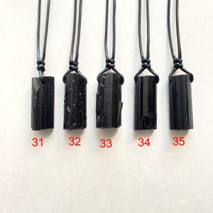 Peut inclure: Cinq pendentifs en cristal de tourmaline noire brute suspendus &agrave; des colliers en cordon noir. Chaque pierre pr&eacute;sente une forme colonnaire allong&eacute;e unique avec des textures naturelles. Les pendentifs sont num&eacute;rot&eacute;s 31, 32, 33, 34 et 35 en rouge.