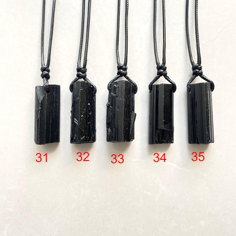 Peut inclure: Cinq pendentifs en cristal de tourmaline noire brute suspendus &agrave; des colliers en cordon noir. Chaque pierre pr&eacute;sente une forme colonnaire allong&eacute;e unique avec des textures naturelles. Les pendentifs sont num&eacute;rot&eacute;s 31, 32, 33, 34 et 35 en rouge.