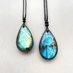 May include: Two teardrop-shaped labradorite pendants with black cords. The stones display iridescent blue, green, and yellow hues. The pendants are likely for necklaces, showcasing natural gemstone jewelry.