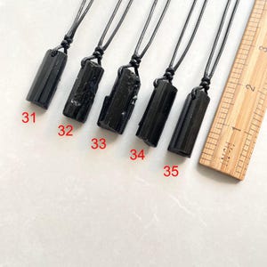 Peut inclure: Cinq pendentifs en pierre noire sur des colliers en cordon noir, &eacute;tiquet&eacute;s de 31 &agrave; 35, dispos&eacute;s &agrave; c&ocirc;t&eacute; d'une r&egrave;gle en bois affichant des mesures en pouces. Chaque pendentif pr&eacute;sente une pierre brute, verticale et cylindrique.