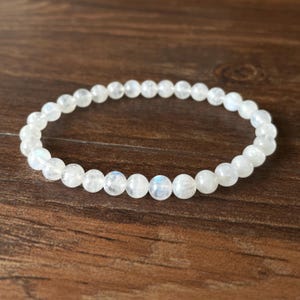 May include: A white beaded bracelet with a subtle iridescent sheen. The bracelet is made of small, round beads strung together on an elastic band. The beads have a smooth texture and a slight glow, set against a dark wood background.