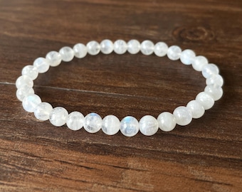 May include: A white beaded bracelet with a subtle iridescent sheen. The bracelet is made of small, round beads strung together on an elastic band. The beads have a smooth texture and a slight glow, set against a dark wood background.