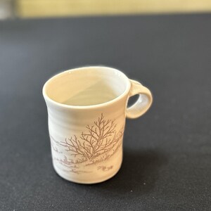 May include: A small, off-white ceramic mug with a brown landscape design featuring a tree. The mug has a curved handle and a slightly flared rim. The design wraps around the mug, creating a rustic aesthetic.