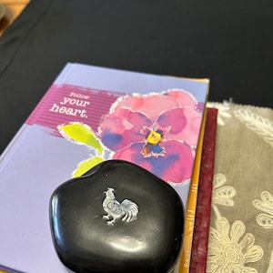 May include: A black, heart-shaped decorative stone with a silver rooster design. The stone is resting on a stack of books, including one with the text "Follow your heart" and a floral illustration. A decorative floral patterned cloth is also visible.