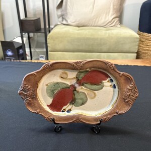 May include: A decorative ceramic serving tray with a brown rim and a cream-colored interior. The tray features a floral design with red and green accents. The tray is supported by a black metal stand, perfect for displaying food or decorative items.