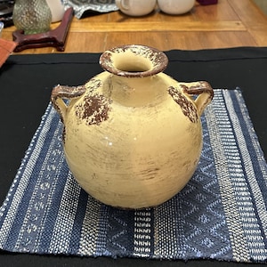 May include: A cream-colored ceramic vase with a round body and two handles. The vase has a distressed finish with brown accents and a wide opening. It sits on a blue and white patterned placemat.