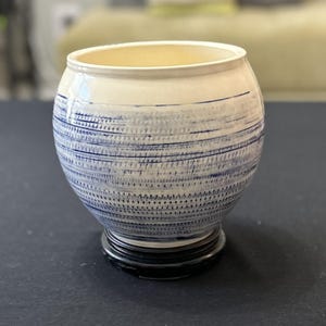 May include: A white ceramic vase with a blue and white striped pattern. The vase has a round body and a black base.