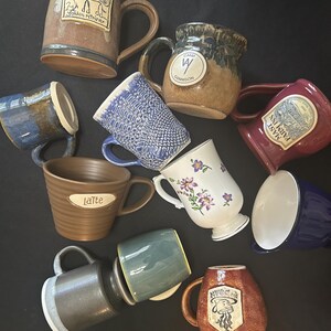 May include: A collection of ten ceramic mugs in various colors, shapes, and finishes. Some mugs feature decorative elements like a golden retriever illustration, the text "Camp Gunnison," "Latte," "Mast Farm Inn," or "The Bean," and floral patterns.