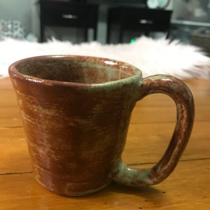 May include: A handmade ceramic mug with a brown and green glaze. The mug has a rounded body and a curved handle. The exterior features a textured, marbled effect. The rim and handle are a muted green color. The mug is sitting on a wooden surface.