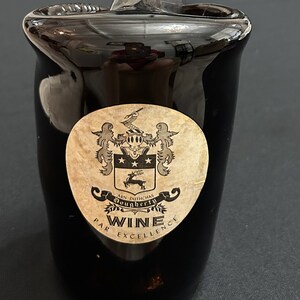 May include: A black glass wine bottle stopper with a vintage-style label featuring a coat of arms and the text "Arn Duthchas Dougherty Wine Par Excellence".