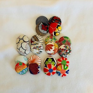 Puede incluir: Una colección de 12 botones redondos de tela con varios patrones coloridos de inspiración japonesa. Los botones están dispuestos en semicírculo sobre una superficie blanca.