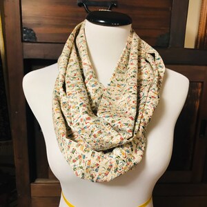 Puede incluir: Una bufanda infinita de color crema con un patrón intrincado y colorido. La bufanda está hecha de una tela suave y tejida y se muestra en un maniquí blanco.