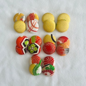 May include: A collection of ten round fabric buttons with various colorful patterns. The buttons feature a mix of red, orange, yellow, green, and white colors. The patterns include floral designs, geometric shapes, and abstract patterns.