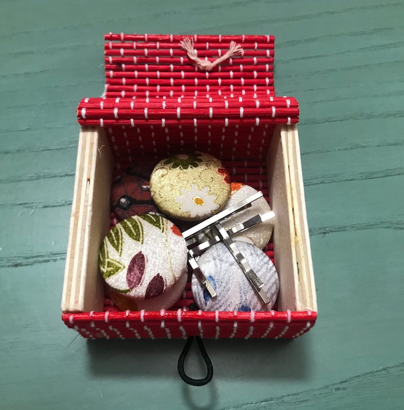 Puede incluir: Una caja con estampado rojo y blanco con una tapa con bisagras. La caja contiene cuatro botones redondos de tela con dise&ntilde;os florales y dos pinzas para el pelo plateadas.