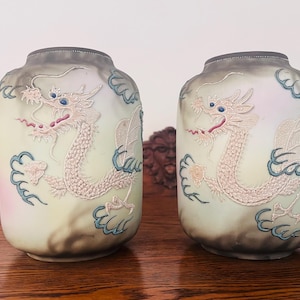 Op de afbeelding: Twee bijpassende keramische vazen met een lichtgroene en grijze gradiënt. Elke vaas heeft een geborduurd drakenontwerp in crème, blauw en roze. De vazen zijn cilindrisch met een licht uitlopende bovenkant.