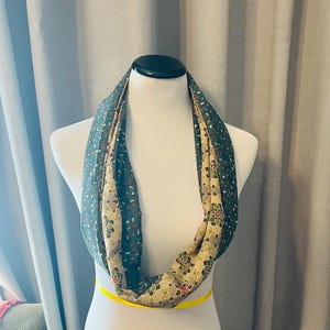 Op de afbeelding: Een groene en beige oneindige sjaal met een bloemenpatroon. De sjaal wordt getoond op een wit mannequin.