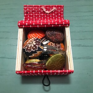 Puede incluir: Una caja con un patr&oacute;n rojo y blanco que contiene una selecci&oacute;n de pinzas para el cabello cubiertas de tela. Las pinzas vienen en una variedad de colores y patrones, incluyendo dise&ntilde;os florales, geom&eacute;tricos y abstractos.