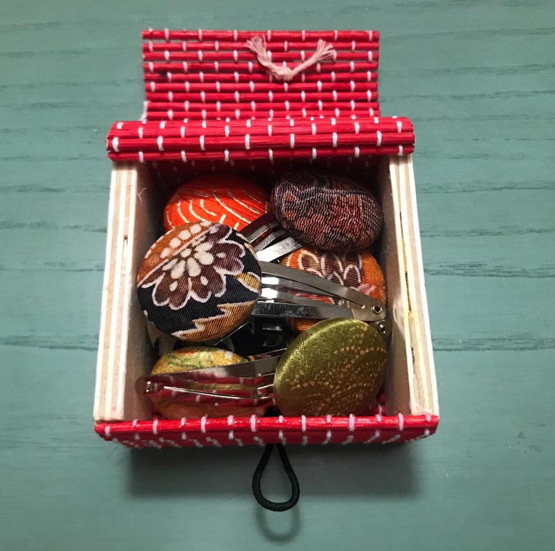 Puede incluir: Una caja con un patr&oacute;n rojo y blanco que contiene una selecci&oacute;n de pinzas para el cabello cubiertas de tela. Las pinzas vienen en una variedad de colores y patrones, incluyendo dise&ntilde;os florales, geom&eacute;tricos y abstractos.