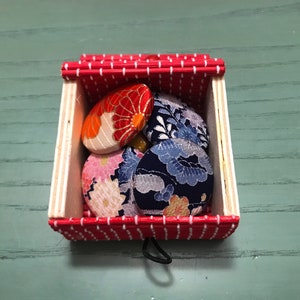 Puede incluir: Una caja de rayas rojas y blancas con una tapa con bisagras contiene cinco botones cubiertos de tela. Los botones est&aacute;n decorados con motivos florales en tonos rojos, azules y blancos.