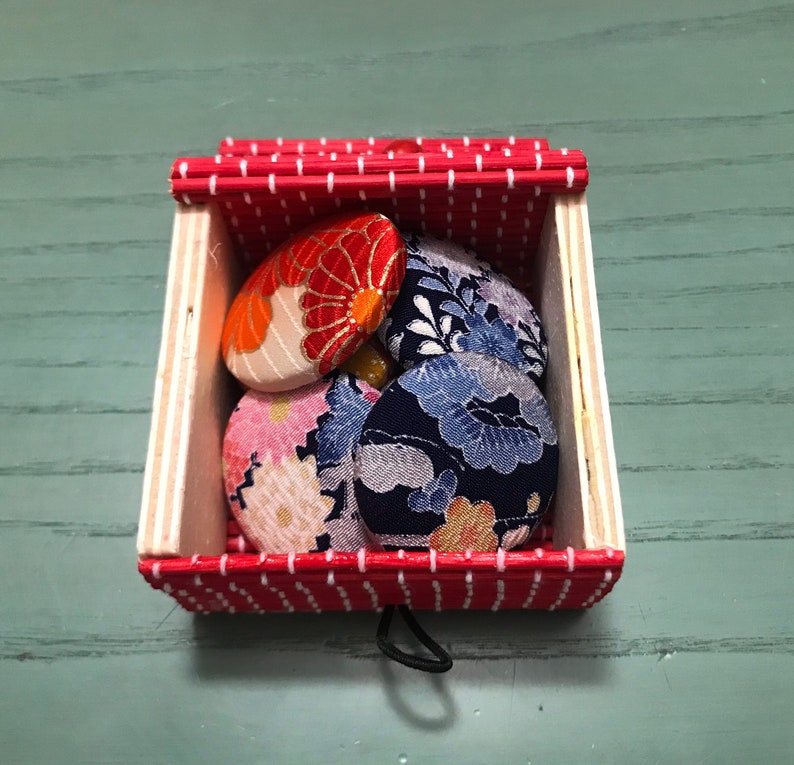 Puede incluir: Una caja de rayas rojas y blancas con una tapa con bisagras contiene cinco botones cubiertos de tela. Los botones est&aacute;n decorados con motivos florales en tonos rojos, azules y blancos.