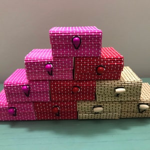 Puede incluir: Una pila de peque&ntilde;as cajas decorativas hechas de bamb&uacute; tejido. Las cajas son de color rosa, rojo y crema, y tienen peque&ntilde;as perillas redondas de colores.