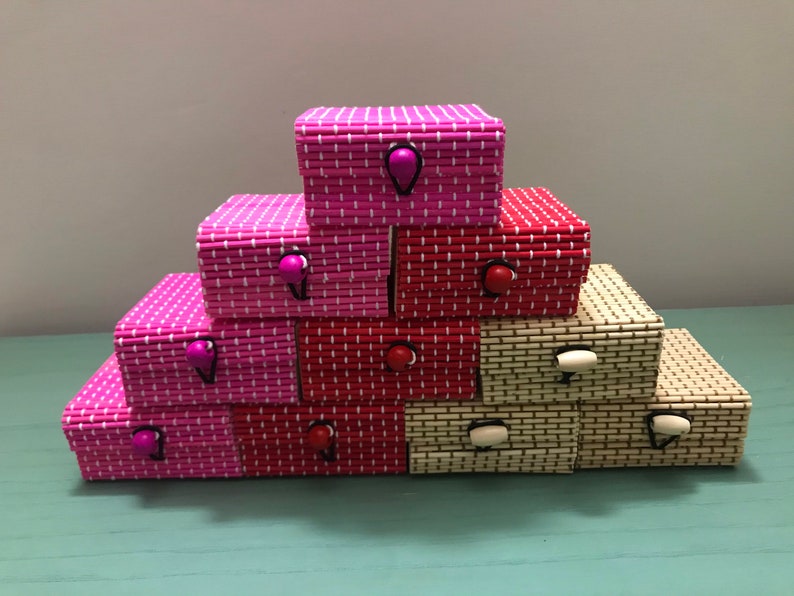 Puede incluir: Una pila de peque&ntilde;as cajas decorativas hechas de bamb&uacute; tejido. Las cajas son de color rosa, rojo y crema, y tienen peque&ntilde;as perillas redondas de colores.
