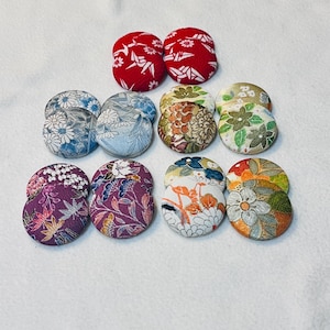 May include: A collection of fabric-covered buttons in various colors and patterns. The buttons feature floral and geometric designs in shades of red, blue, purple, green, and orange. The buttons are round and appear to be handmade.